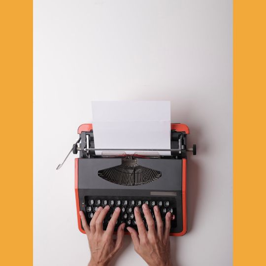 Fotografia de cima de um par de mãos digitando em uma máquina de escrever de bordas alarannjadas sobre um fundo branco